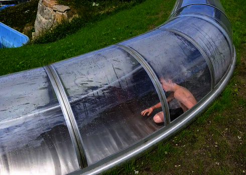 Kind rutscht in transparenter Röhrenrutsche aus Edelstahl über grüne Wiese.