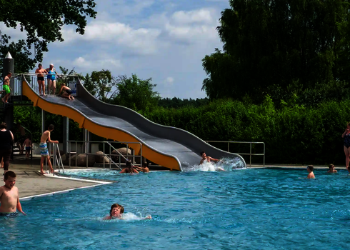 Ein Freibad mit Badgästen im Wasser und an Land, im Hintergrund eine gelbe Breitwellenrutsche.