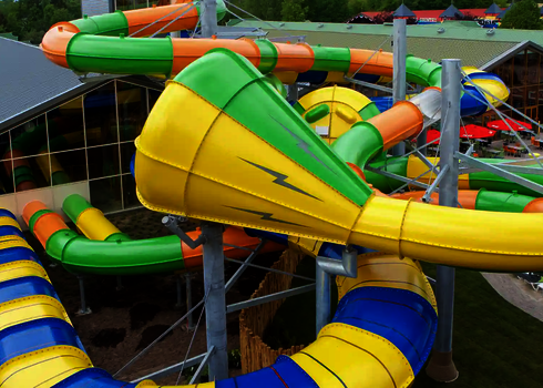 Grün-gelbe Crazy Cone Rutsche mit Blitzsymbol, Teil einer großen Rutschenanlage vor Glasfassade.