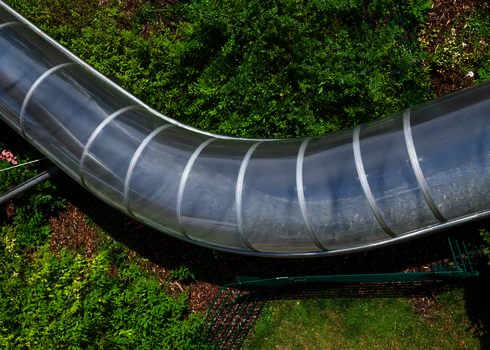 Blick von oben auf eine transparente Röhrenrutsche in grüner Umgebung.