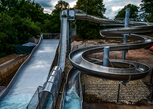 Unterschiedliche Wasserrutschen aus Edelstahl mit Auslaufbecken vor einer Waldumgebung.