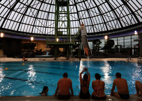 Mehrere Personen sitzen am Beckenrand eines Hallenbades und schauen auf eine weitere Person, die die Fallrutsche herunter rutscht.