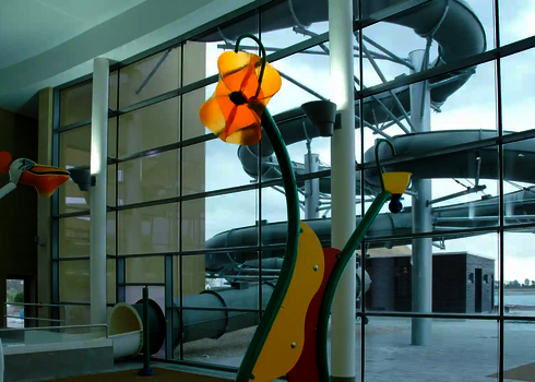 Eine wassersprühende Blume im Schwimmbad, im Hintergrund eine große graue Wasserrutsche im Außenbereich.