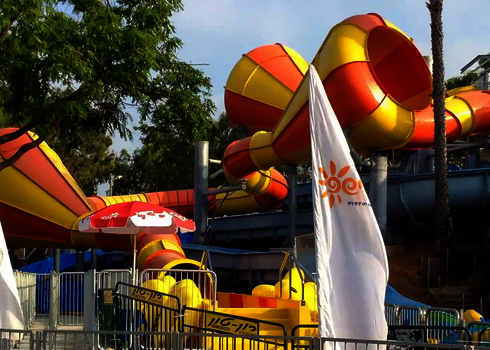 Zwei Crazy Cones mit gelb-oranger Röhre in einer Wasserpark-Anlage hinter Sonnenschirmen.