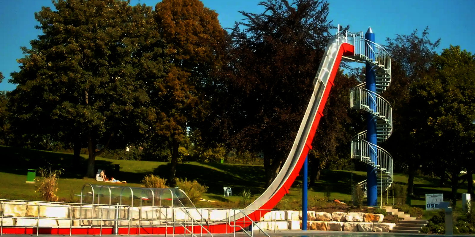 Freifallrutsche mit rot-weißem Design mündet in einem Auslaufbecken, in einem Freibad mit Schwimmern.