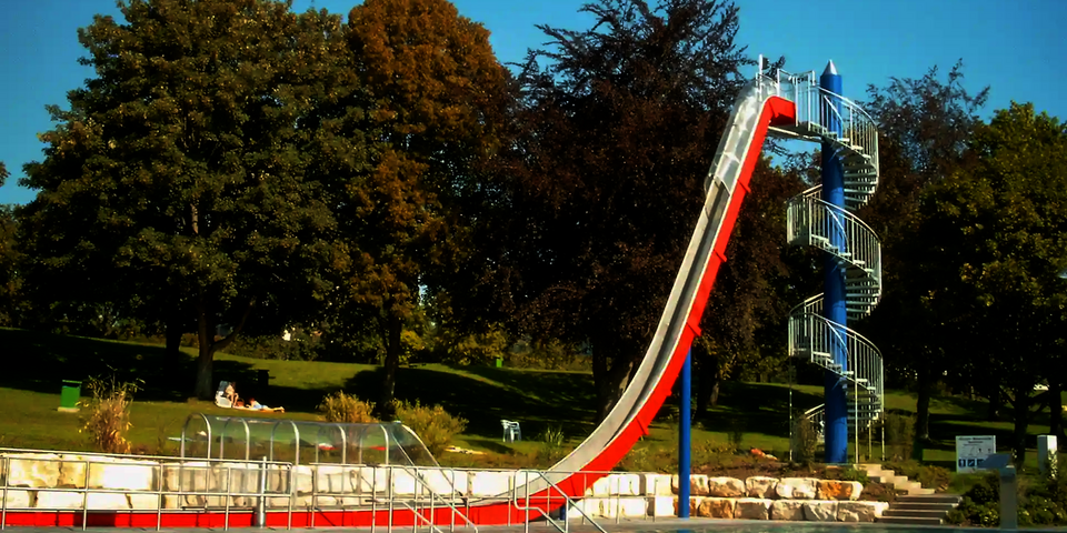 Freifallrutsche mit rot-weißem Design mündet in einem Auslaufbecken, in einem Freibad mit Schwimmern.