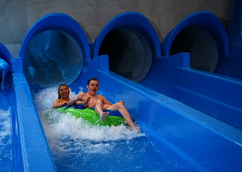 Zwei Personen rutschen auf einem grünen Reifen aus einer blauen Wasserrutsche.