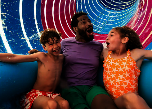 Ein Mann rutscht mit zwei Kindern im Rutschreifen durch eine farbig beleuchtete Röhrenrutsche.