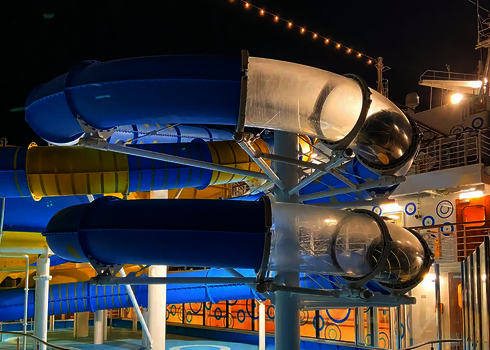 Eine blaue und gelbe RR1200 Wasserrutsche mit transparenten Acryl-Abschnitt auf einem Kreuzfahrtschiff bei Nacht.