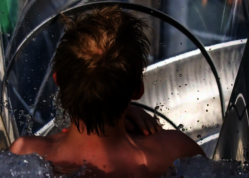 Ein Junge rutscht in einer transparenten Freifallrutsche aus Edelstahl ins Wasser.