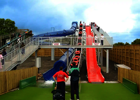 Blick auf zwei Wasserrutschen, in der Mitte ein Reifenlift, Personen stehen davor und schauen zu.