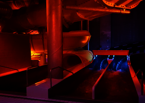 Zwei gelbe Röhrenrutschen mit Wasserlauf leuchten orange-blau in einem dunklen Hallenbad.