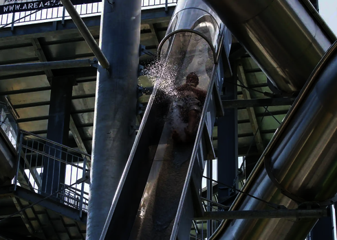 Person rutscht mit Wasserfontäne durch eine steile, transparente Edelstahlrutsche.
