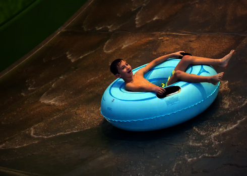 Ein Junge rutscht lachend mit einem blauen Reifen auf der Sidewinder in einem Hallenbad.