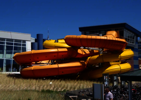 Gelbe und orangefarbene Family Twister Wasserrutsche vor einem Freizeitbad.