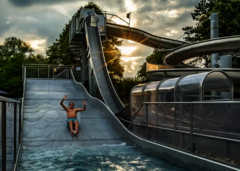 Eine Person rutscht mit erhobenen Händen eine Breitwellenrutsche herunter, daneben befinden sich weitere Wasserrutschen aus Edelstahl.