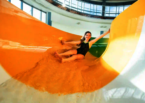 Mädchen rutscht lachend eine bunte Indoor-Wasserrutsche in orangefarbenem Bereich hinunter.