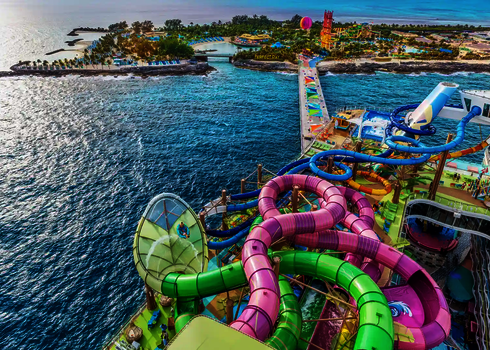 Blick von oben auf eine farbenfrohe Rutschenanlage an Deck eines Kreuzfahrtschiffes, im Hintergrund eine Insel.