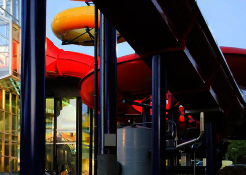 Mehrere bunte Röhrenrutschen verlaufen außen an einem Glasgebäude entlang.