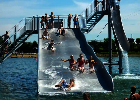 Kinder rutschen lachend nebeneinander eine Breitwellen Wasserrutsche im Freibad herunter, daneben befindet sich eine Fallrutsche.