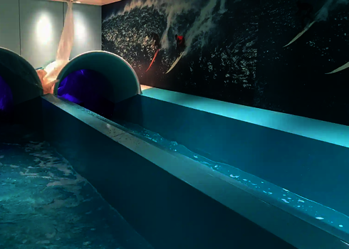 Two brightly coloured slide ends with a dividing bar in a turquoise pool, with a picture of a surfer in the background.