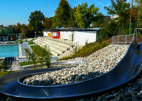Eine Wasserrutsche die sich einen Hang mit Steinen herunterschlängelt und in ein Schwimmbecken mündet.