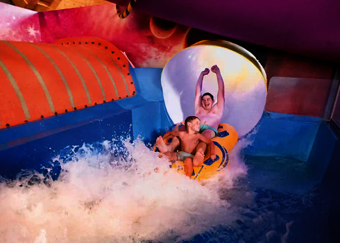 Zwei Personen rutschen lachend aus einer bunten Wasserrutsche in ein blaues Auslaufbecken.