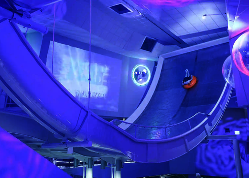 Person auf Reifen rutscht die beleuchtete Boomerang-Wand hinunter, alles in Blau-Violett.