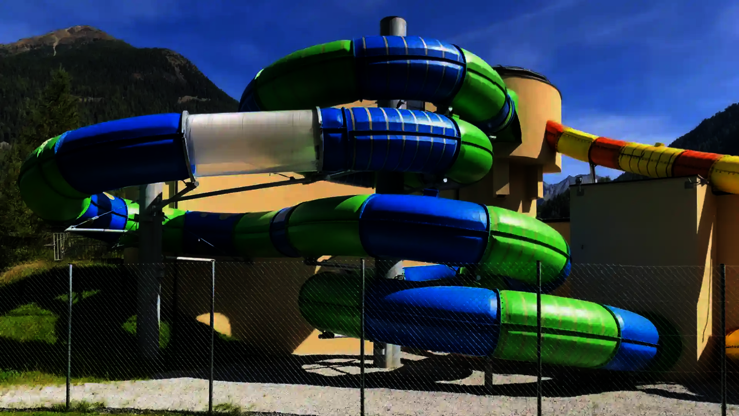 Farbige gedämmte Wasserrutsche in Blau und Grün mit gelbem Startstück vor Bergkulisse.