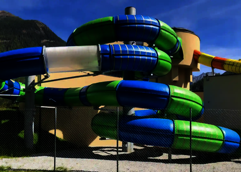 Farbige gedämmte Wasserrutsche in Blau und Grün mit gelbem Startstück vor Bergkulisse.