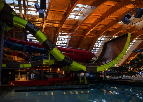Gelb-schwarze Boomerang-Rutsche in einem Wasserpark mit Wasserbecken, Palmen und bunter Kulisse.