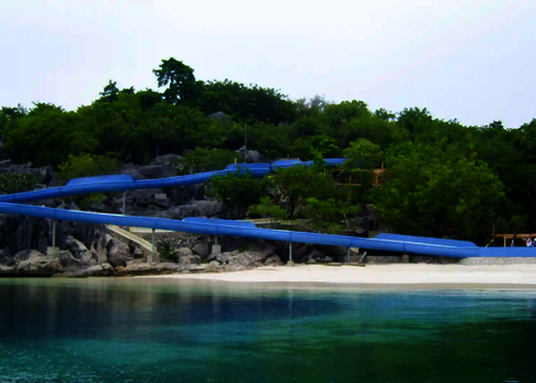 Lange blaue Wasserrutsche führt entlang Felsen durch tropische Umgebung bis an einen Sandstrand.