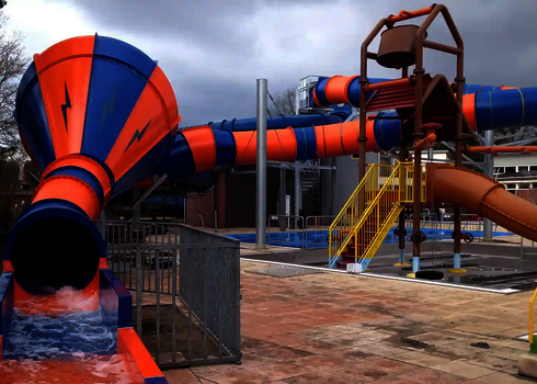 Crazy Cone mit orange-blauer Rutsche neben einem Wasserspielplatz mit gelber Treppe.