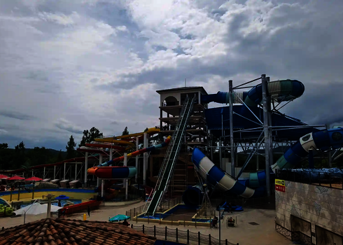 Mehrere bunte Rutschen und ein zentraler grüner Reifenlift in einem Wasserpark mit Hügelkulisse.