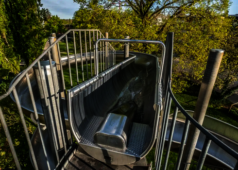 Blick von oben auf den Einstieg einer Edelstahlrutsche mit Wasserlauf in grüner Parkanlage.
