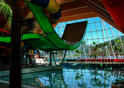 Grüne Boomerang-Rutsche über Innenbecken, im Hintergrund große Glasfassade und Palmen.