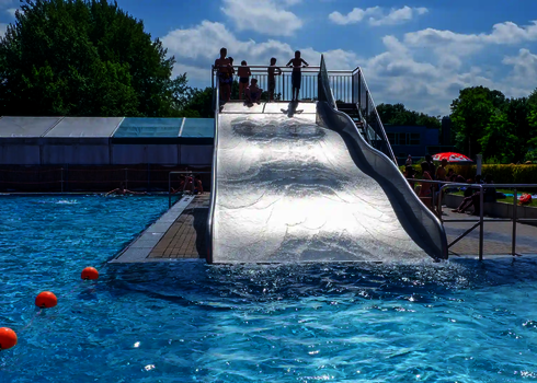 Oben auf einer Breitwellenrutsche stehen Kinder, die auf das Schwimmbecken schauen.