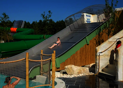 Ein Kind rutscht eine Breitwellenrutsche herunter, im Hintergrund befindet sich eine grünen Röhrenrutsche.