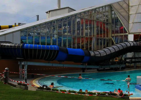 Blick auf den Außenbereich eines Schwimmbades mit einem Pool und einer schwarz-blauen Rutsche.