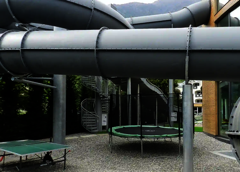 Eine graue RR1200 Wasserrutsche verläuft oberhalb eines Outdoor-Bereichs mit Tischtennisplatte und Trampolin.