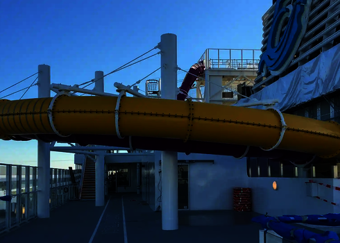 Gelbe Röhrenrutsche auf einem Kreuzfahrtschiff, getragen von Stützen und mit Tageslichteffekten.