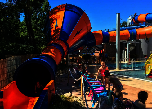 Kind verlässt orange-blaue Crazy Cone Rutsche, im Hintergrund ein Wasserspielplatz.