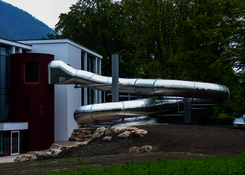 Edelstahlrutsche, die an ein Gebäude grenzt, vor wäldlichem Hintergrund.