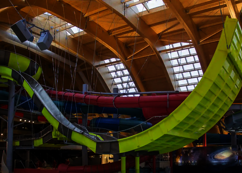Überdachtes Hallenbad mit unterschiedlichen bunten Wasserrutschen, im Vordergrund eine gelbe Boomerang-Rutsche.