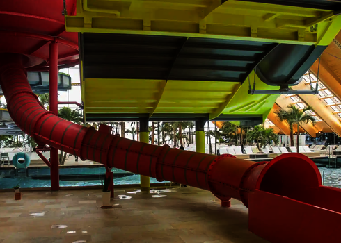 Innenansicht eines Hallenbas mit einer roten Röhrenrutsche im Vordergrund und einer gelb-grauen Wasserrutsche daneben.