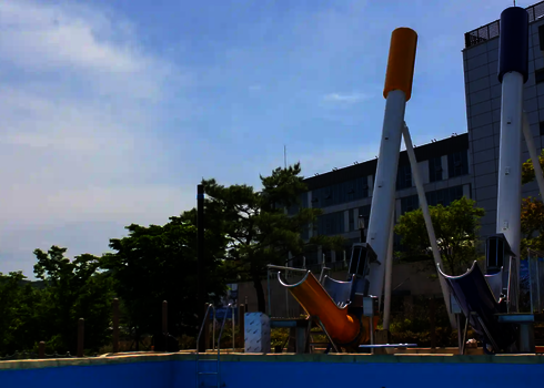 Ein gelber und ein blauer Cannonball vor einem Schwimmbecken, blauer Himmel und Häuser im Hintergrund.