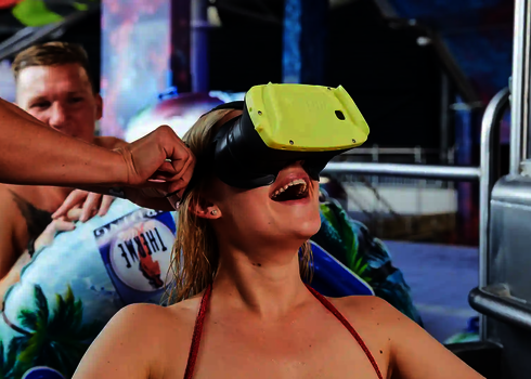 Frau trägt lachend eine VR-Brille, im Hintergrund sitzt ein Mann.