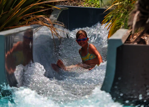 Ein Mädchen rutscht lachend durch den Wildwasserfluss mit Bepflanzung an der Seite.