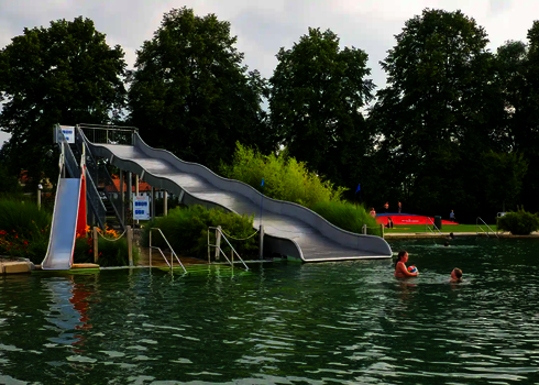 Freibad mit einer Breitwellenrutsche und einer kleineren Wasserrutsche daneben, im Schwimmbecken befinden sich Badegäste.