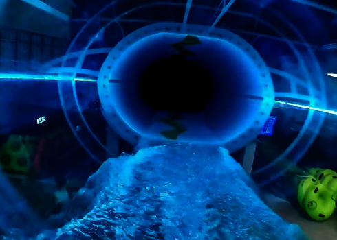 Blick von innen durch ein beleuchtetes Acrylrutschen-Element auf einen dunklen Tunnel.